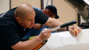 A man examining a car's dent closely with a flashlight and tools, another person in the background, inside a Paintless Dent Repair Training Academy workshop.