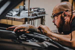 A bald tattooed man wearing glasses performs paintless dent repair on a car's body with precision tools in a workshop.