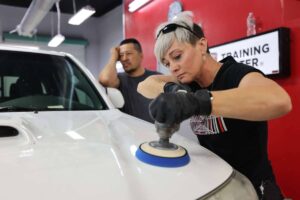 A woman uses a power buffer for paint correction on a car hood while a man observes in a training center.