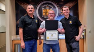 Three men standing in an office, with the middle one holding a certificate; they are all smiling and dressed in black polo shirts, with a logo labeled "TOTAL Recon" in the background.