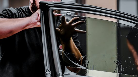 A person's hands visible through a shattered car window with tinting, with another hand holding the frame from outside.