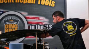 A man in a black "ding king" shirt works on a car in a workshop with a sign saying "PDR Training" in the background.