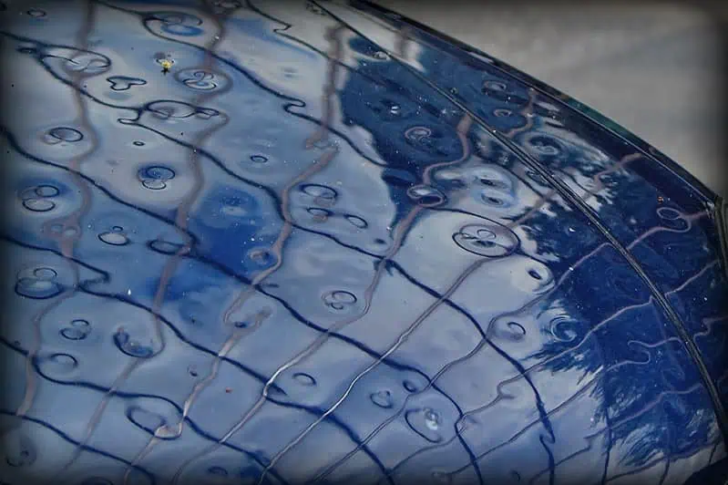 Close-up of a blue car hood reflecting hailstones, creating a patterned effect with subtle tree reflections in the background.