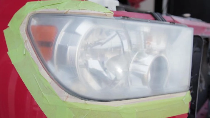 Car headlight covered with green tape, possibly for renewal or painting, set against a blurry red background.