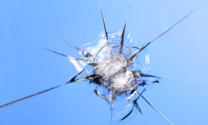 A close-up image of a shattered windshield with sharp fragments radiating outward against a clear blue sky background.